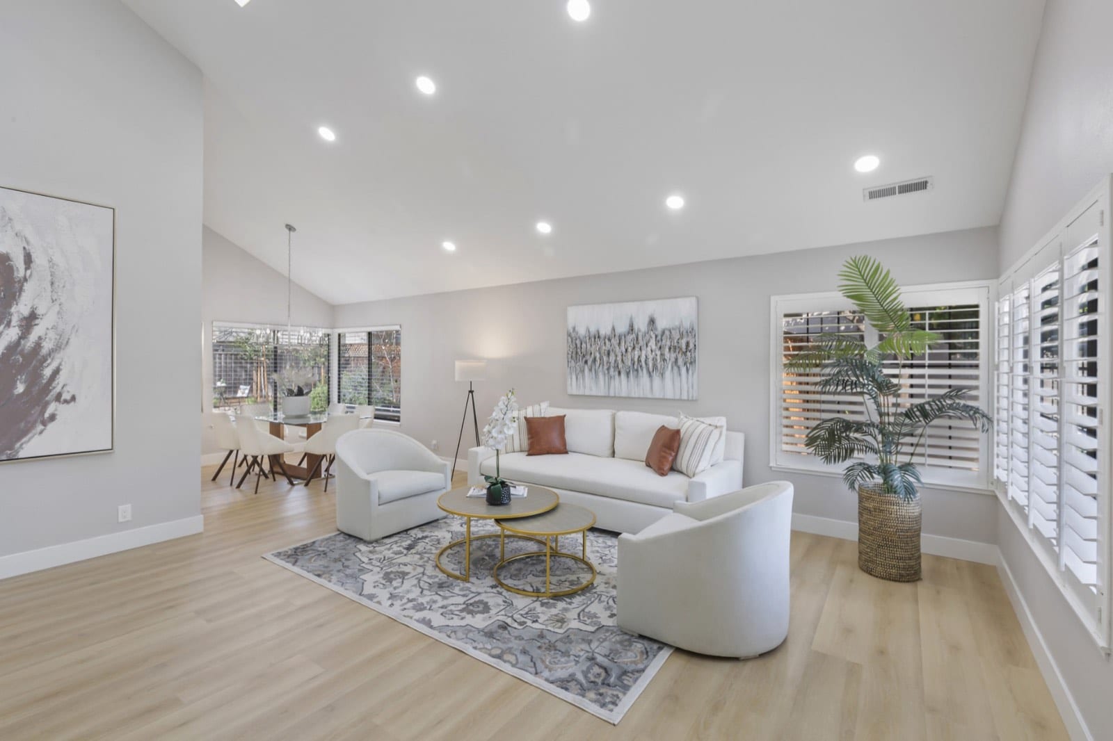 Vaulted living room with white sofa and chairs, gold nesting tables, plantation shutters and a view to the dining room