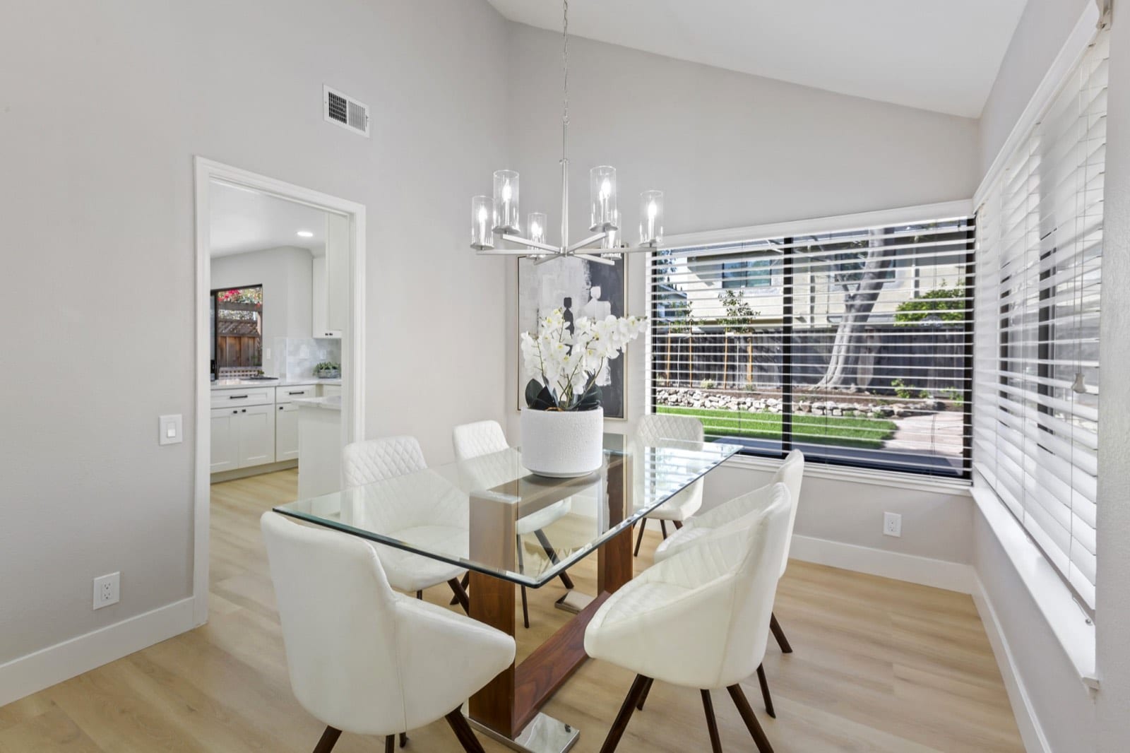 Dining room with a glass table, white chairs, a linear chandelier and a wide window onto the backyard