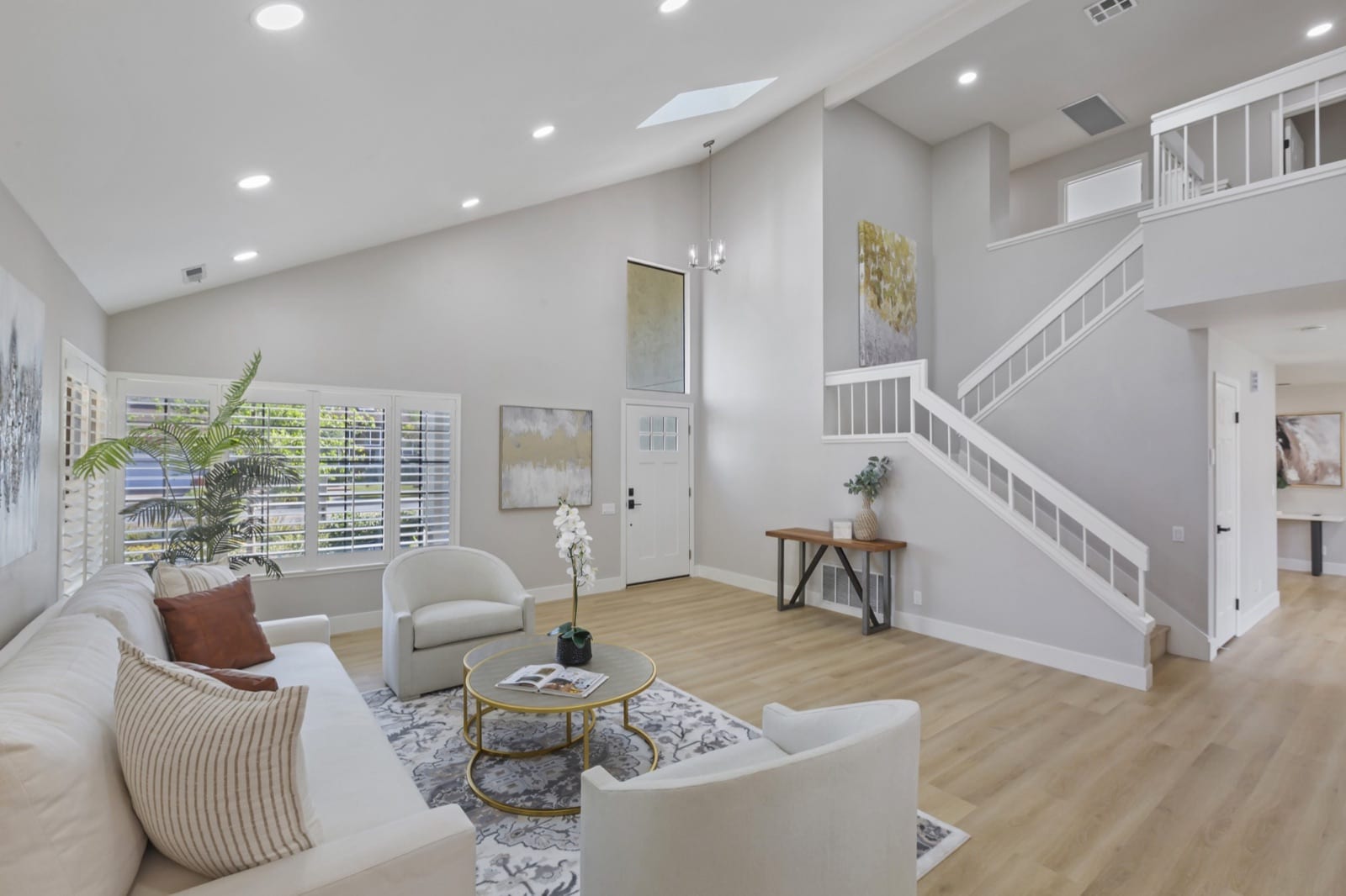 After: staged living room with light plank flooring, white stair rails, soft gray walls and recessed lighting