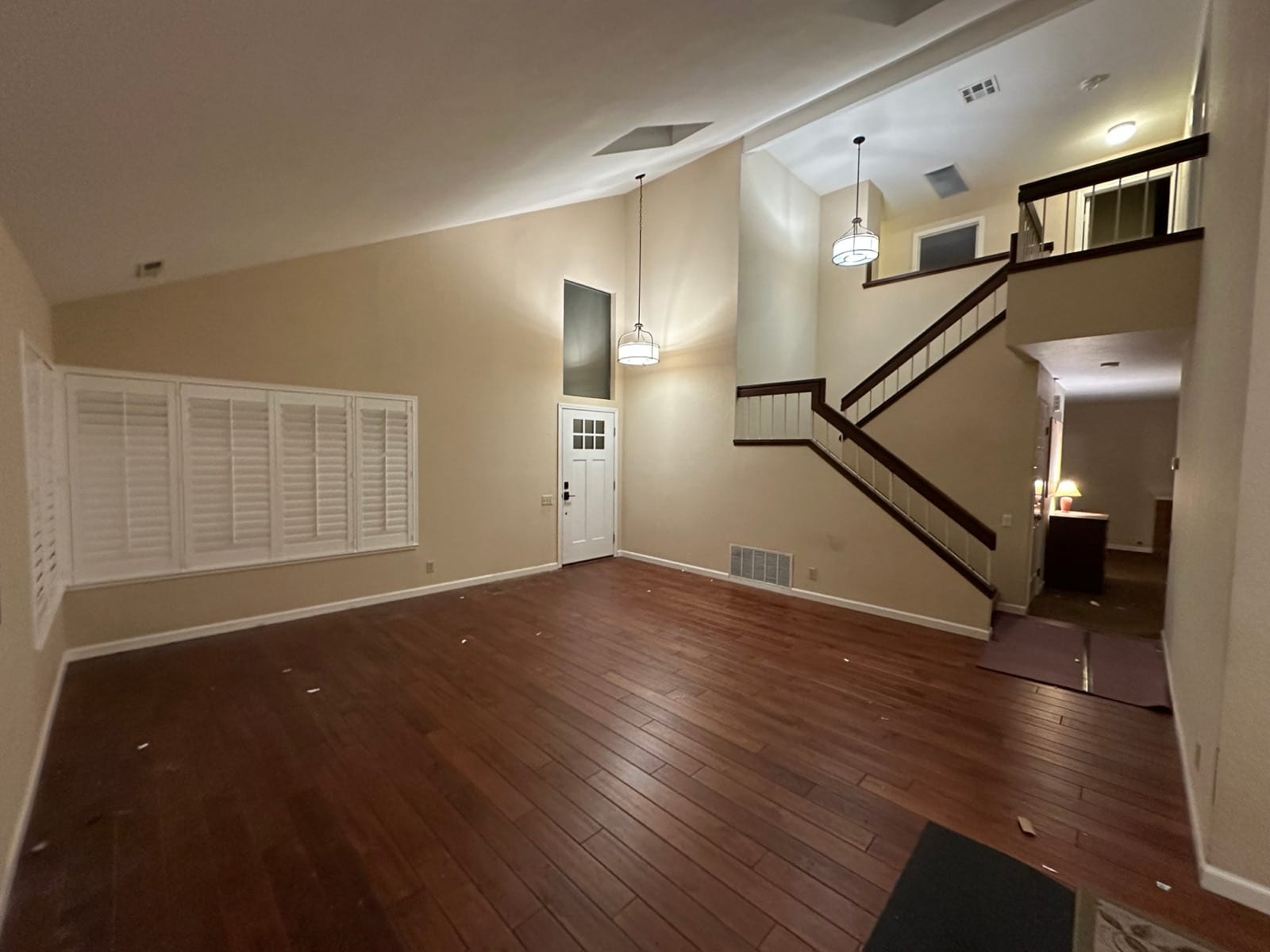 Before: empty living room with dark wood floors, beige walls, dark stair rails and pendant lights under a vaulted ceiling