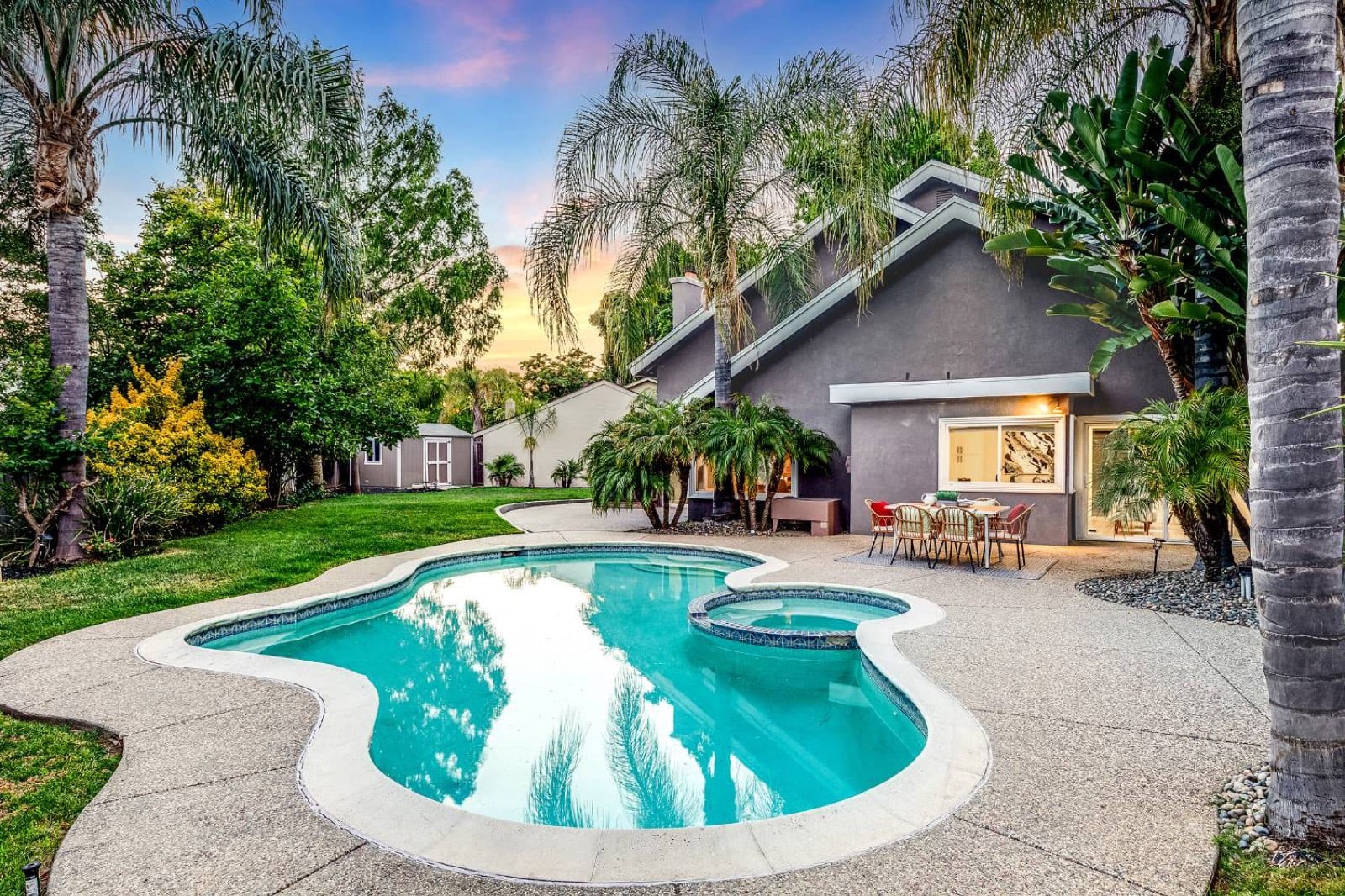 Backyard at dusk: freeform pool and spa ringed by palms, with a patio dining set